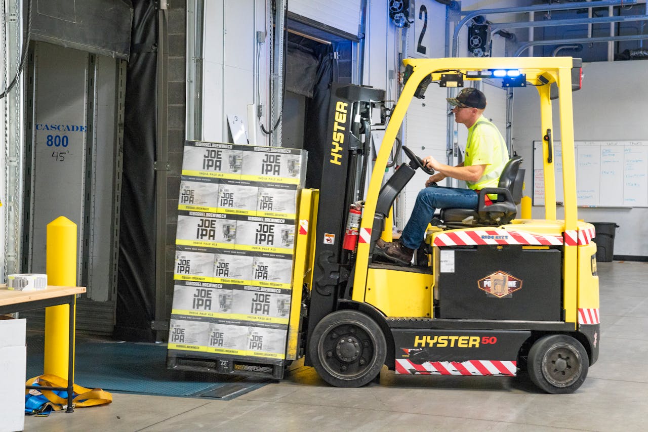 About A worker drives a Hyster forklift moving Joe IPA boxes in a warehouse.